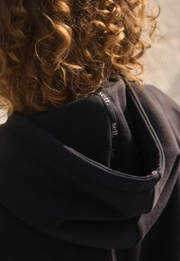 Schwarzer Kapuzenpullover aus strukturiertem, weichem Stoff. Das Innere der Kapuze zeigt ein dezentes Druckdetail mit dem Wort "selbst." Lockige Haare im Hintergrund.