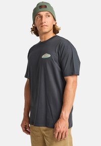 Camiseta de algodón gris oscuro de manga corta, con un pequeño logo verde y rojo en el pecho, combinada con pantalones de pana marrón claro.