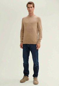 Beiger Strickpullover mit Rundhalsausschnitt, langen Ärmeln, kombiniert mit dunkelblauen Jeans und hellbraunen Sneakers. Minimalistisches Design und Textur.