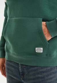 Persona che indossa una felpa verde con tasca frontale e piccolo etichetta bianca "CREEKS", mano appoggiata dentro la tasca, abbinata a jeans blu.