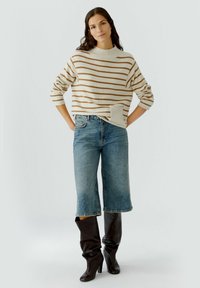 Cremfarbener gestreifter Pullover mit hohem Ausschnitt, kombiniert mit hellblauen, cropped Jeans. Das Modell trägt braune kniehohe Stiefel.