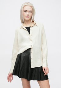 Camisa bege com botões, tecido texturizado, mangas compridas e gola, combinada com uma minissaia preta em pele com corte evasê.