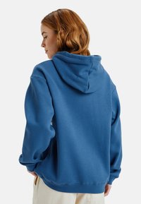 Sudadera azul hecha de tejido suave, con capucha ajustable, puños de canalé y un corte relajado. Sin logotipos ni patrones visibles.