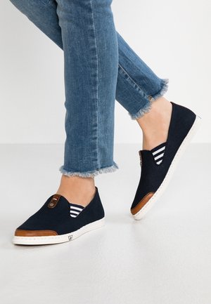Personne portant un jean bleu à ourlet effiloché et des chaussures décontractées noires sans lacets avec des accents marron sur les orteils et des côtés élastiques rayés blancs.