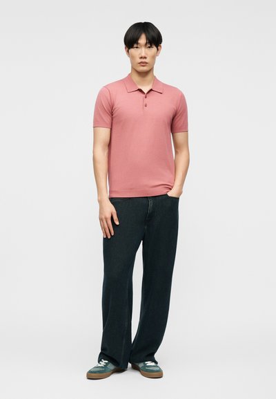 Νεαρός άνδρας που φοράει μια κοντομάνικη πόλο μπλούζα σε ξεθωριασμένο ροζ χρώμα, φαρδιά σκούρα τζιν και πράσινα αθλητικά παπούτσια, στέκεται με το ένα χέρι στην τσέπη.