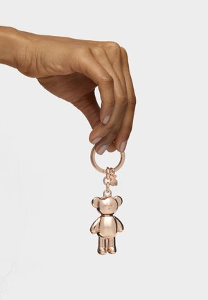 Porte-clés en forme d'ours en peluche en or rose tenu par une main. Le porte-clés présente une surface polie avec un corps et des oreilles arrondis, attaché à un anneau circulaire.