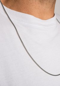 Metalliketju kaulaketju, hopeanvärinen, jossa on sileä, kiiltävä pinta ja tasaiset lenkit. Päälle on puettu valkoinen puuvillapaita.