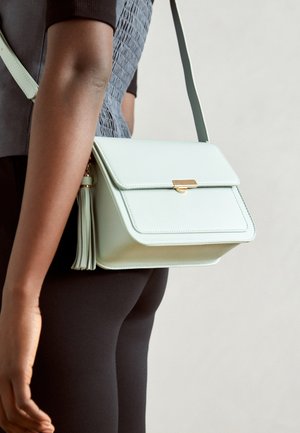 Persona con ropa negra ajustada lleva un bolso bandolera azul claro con cierre dorado y detalle de borla en el lateral.