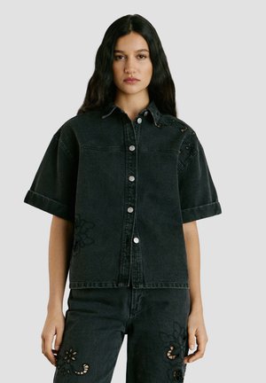 Frau mit langen dunklen Haaren, die ein schwarzes, kurzärmliges Denim-Hemd mit Knöpfen und passende Hosen mit floraler Cutout-Stickerei trägt.