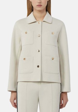 Vrouw draagt een crèmekleurig jasje met vier voorzakken en gouden vierkante knopen, gecombineerd met bijpassende crèmekleurige broek met zwarte zijstrepen.