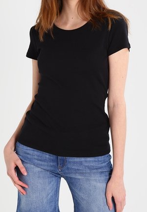 Woman wearing a plain black short-sleeve t-shirt and blue jeans, standing with one hand on her hip against a white background.