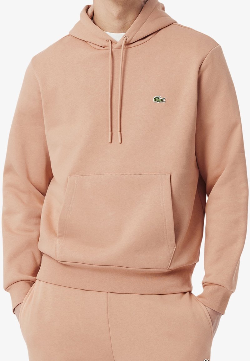 Sweat à capuche rose clair avec poche kangourou à l'avant et cordons, avec un petit logo de crocodile vert sur la poitrine gauche.
