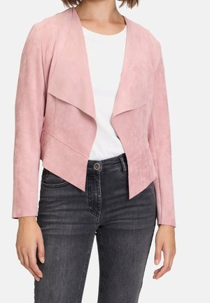 Femme portant une veste en suède rose clair ouverte sur le devant, par-dessus une chemise blanche et un jean skinny noir délavé.