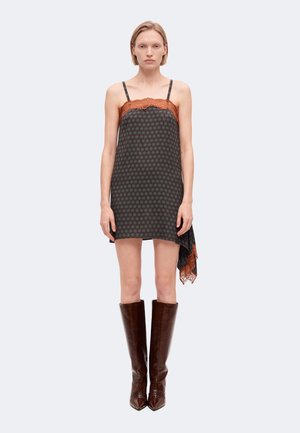 Femme portant une robe nuisette sombre à motifs avec une bordure en dentelle orange sur le haut et l'ourlet, associée à de grandes bottes en cuir marron, debout sur un fond blanc.
