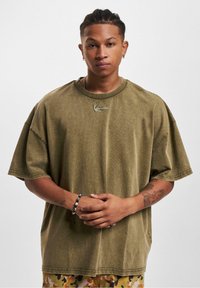 Olive green oversized cotton t-shirt with short sleeves, ribbed collar, and a small embroidered detail on the chest. Smooth texture.