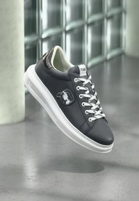 Αθλητικό παπούτσι με επάνω μέρος από σκούρο ναυτικό δέρμα, λευκή σόλα και αντίθετα κορδόνια. Διακριτικό λογότυπο Karl Lagerfeld στο πλάι και υφασμένες λεπτομέρειες στο πίσω μέρος.