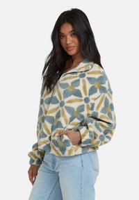 Polarowy sweter z kwiatowym wzorem w odcieniach niebieskiego, żółtego i kremowego. Posiada zamek błyskawiczny do połowy, wysoki kołnierz oraz kieszenie z przodu. Miękka tekstura.