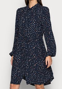Abito lungo a maniche lunghe blu navy con colletto, chiusura a bottoni frontale, cintura in vita e macchie astratte sparse in azzurro chiaro e beige.