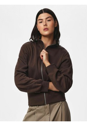Vrouw met steil donker haar die een dichtgeritst donkerbruin jasje en beige broek draagt, poseert met één hand bij haar kraag.