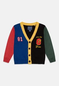 Spalvingas megztinis kardiganas, pasižymintis mėlynomis, juodomis, raudonomis ir žaliomis dalimis, geltonu V formos kaklu, sagomis ir siuvinėjimais, tokiais kaip "81" ir "G Guess."