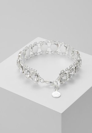 Zilveren kettingarmband met heldere ronde edelstenen en een kleine ronde metalen bedel op een lichtgrijze sokkel.