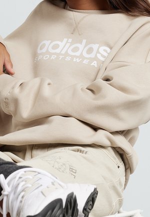 Person trägt beige Adidas-Sportbekleidung, Sweatshirt und Hose, mit weißen Sneakern, sitzt mit verschränkten Armen und entspannten Beinen.