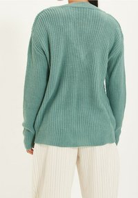 Pull en maille texturée vert clair avec manches longues et coupe décontractée. Ourlet et poignets côtelés, porté sur un pantalon crème à fines rayures.