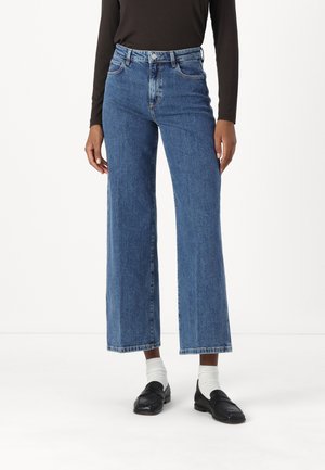 Jeans in denim blu a gamba larga con vita alta e cinque tasche. Presentano un orlo pulito e una texture leggermente sbiadita. Abbinati a mocassini neri.