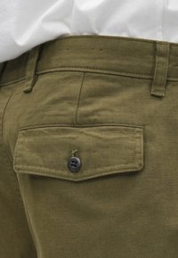 Παντελόνι χρώματος ελιάς με υφή ψαροκόκκαλου, με πίσω τσέπη κλειστή με μαύρο κουμπί και κλ�ασικό σχέδιο στη μέση.