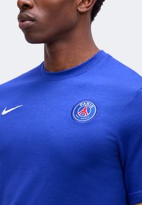 Άνδρας που φοράει μπλε μπλούζα της Paris Saint-Germain με λογότυπο Nike, ορατός από τον λαιμό μέχρι το μέσο του στήθους, μπροστά σε απλό ανοιχτό φόντο.