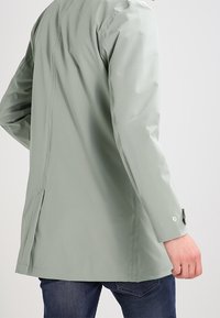 Person trägt eine hellgraue Langarmjacke und blaue Jeans, von hinten vor einem schlichten Hintergrund gezeigt.