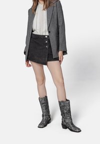 Mujer que lleva un blazer gris, una blusa blanca con encaje, una falda envolvente negra abotonada y botas de cuero grises hasta medio pantorrilla con detalles de tacón y correa.