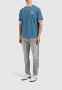 Blauwe katoenen T-shirt met korte mouwen, voorzien van een klein wit logo op de borst, gecombineerd met lichtgrijze jeans en witte sneakers.