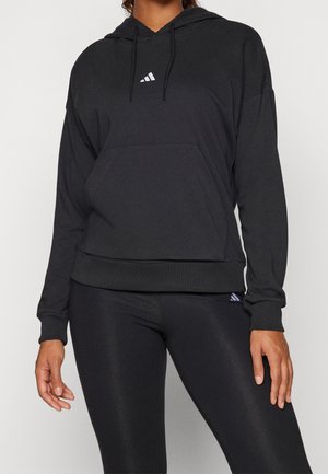 Person trägt schwarzen Hoodie mit kleinem weißem Adidas-Logo und schwarzen Leggings, steht vor einem einfachen hellen Hintergrund.