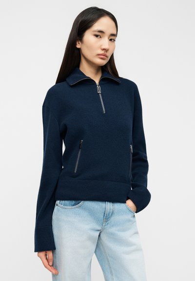 Pull-over bleu marine en tissu doux, avec une fermeture éclair à l'avant, des poches latérales, des manches longues et une coupe décontractée. Texture lisse, design minimaliste.