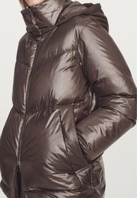 Brązowa kurtka puchowa z połyskującym, pikowanym wzorem, zamek błyskawiczny, wysoki kołnierz i regulowany kaptur. Posiada elastyczne mankiety i boczne kieszenie.
