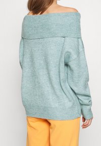 Světlomodrý, nadměrně velký pletený svetr se širokým žebrovaným límcem spadajícím z ramen a dlouhými rukávy. Jemná textura a mírně volný střih.