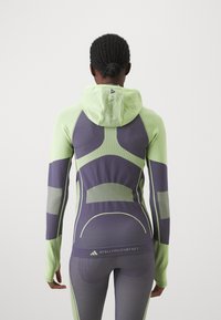 Person i en åtsittande grön och lila huva idrottströja och leggings med geometriska mönster, visad bakifrån.