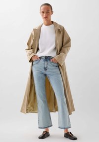 Beige trench coat přes bílou košili, rozšířené světle modré džíny s třásněmi na lemu a černé baleríny. Hladká tkanina a volný střih.