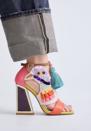 Sandalia de tacón alto con diseño de pájaro colorido, detalle de borlas y correa en el tobillo, usada con vaqueros azules remangados.