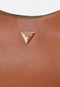 Brązowa torba skórzana o fakturze z trójkątnym metalowym logo z napisem "GUESS U.S.A." na środku. Gładkie szwy wzdłuż krawędzi.