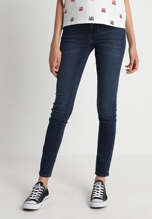 Schmale dunkelblaue Jeans, kombiniert mit schwarzen und weißen Sneakers sowie einem weißen Oberteil mit rotem Schriftzug, gezeigt von der Taille bis zu den Füßen.