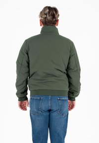 Giacca imbottita verde con collo alto, polsini e vita a costine; abbinata a jeans blu; dettaglio di cucitura visibile sulle maniche.