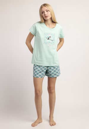 Joven con camiseta de manga corta color verde menta con estampado de sirenas y pantalones cortos a juego, de pie descalza contra un fondo claro.