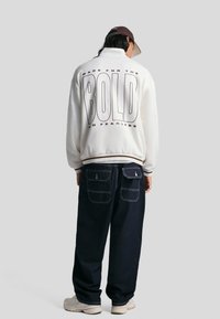 Bela flis jakna z besedilom "BOLD" na hrbtu, v kombinaciji s temnimi jeans hlačami, ki imajo žepne obloge ter belo šivanje, in svetlimi športnimi čevlji.