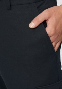 Zwarte broek van een soepele, elastische stof. Pocketdetail zichtbaar met een hand die aan de zijkant rust. Eenvoudig, functioneel ontwerp.