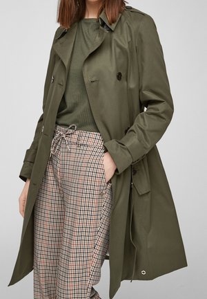 Osoba nosící tmavě zelený trenčkot, sladěný zelený žebrovaný top a vysoké kalhoty s kšiltem v károvaném vzoru se stahovací šňůrkou, s rukama částečně v kapsách.