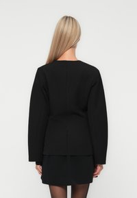 Kvinna med långt blont hår, klädd i en åtsittande svart blazer och matchande kjol, stående mot en enkel vit bakgrund, vänd med ryggen mot kameran.