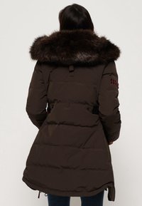 Brun polstret jakke med stor, pelsforet krave. Har bælte i taljen, lommer med lynlås og en mid-længde design. Set bagfra.