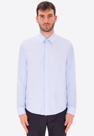 Uomo con la barba che indossa una camicia azzurro chiaro a maniche lunghe e pantaloni scuri, in piedi davanti a uno sfondo bianco uniforme.